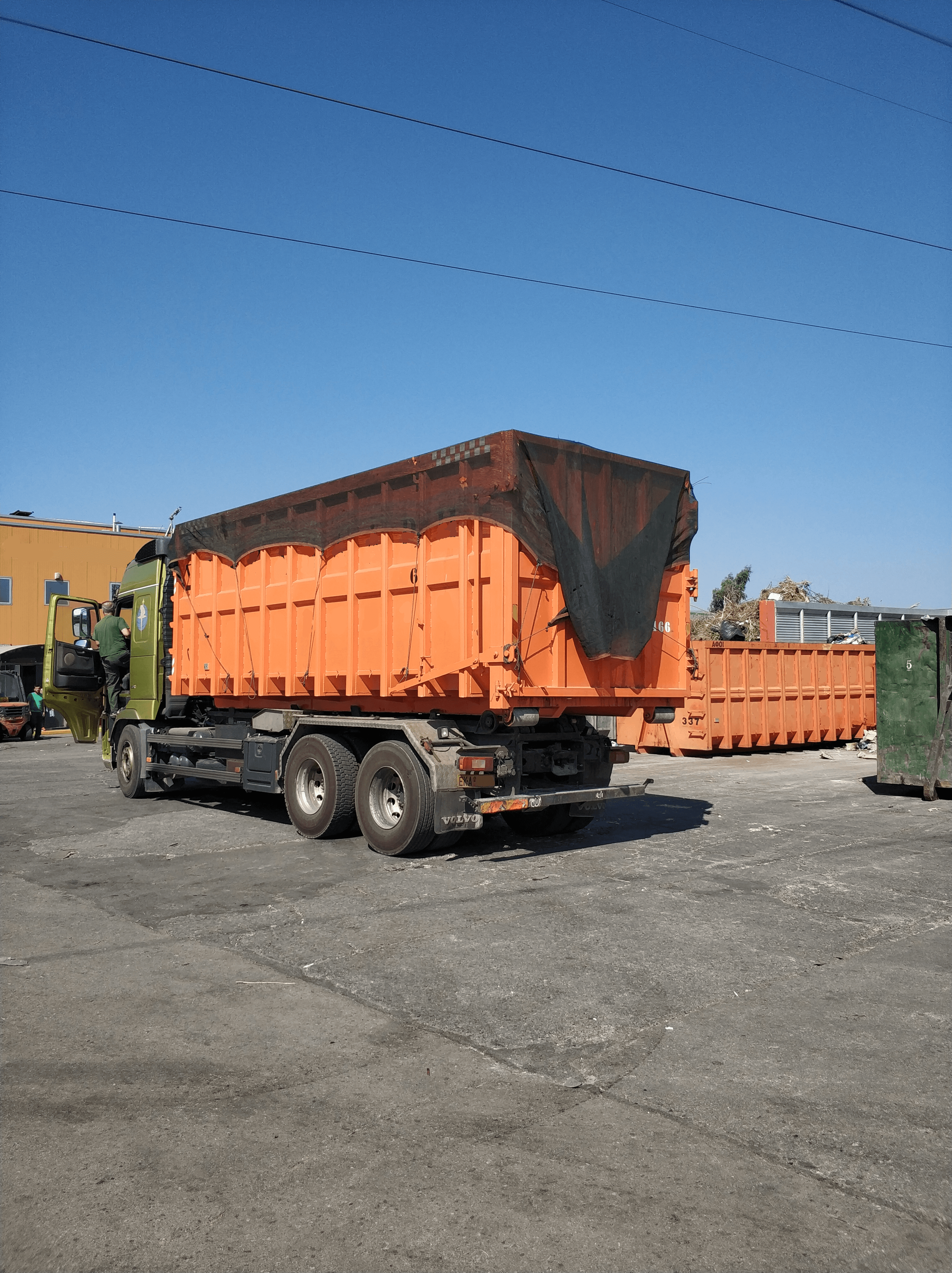 Green Volvo truck with a large orange waste container covered by a black mesh tarp.